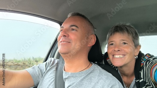 Young adult couple sitting in car, he in the driver's seat looking straight ahead, she in the back seat, leaning between the two front seats, looking out the front window. Traveling with friends. 