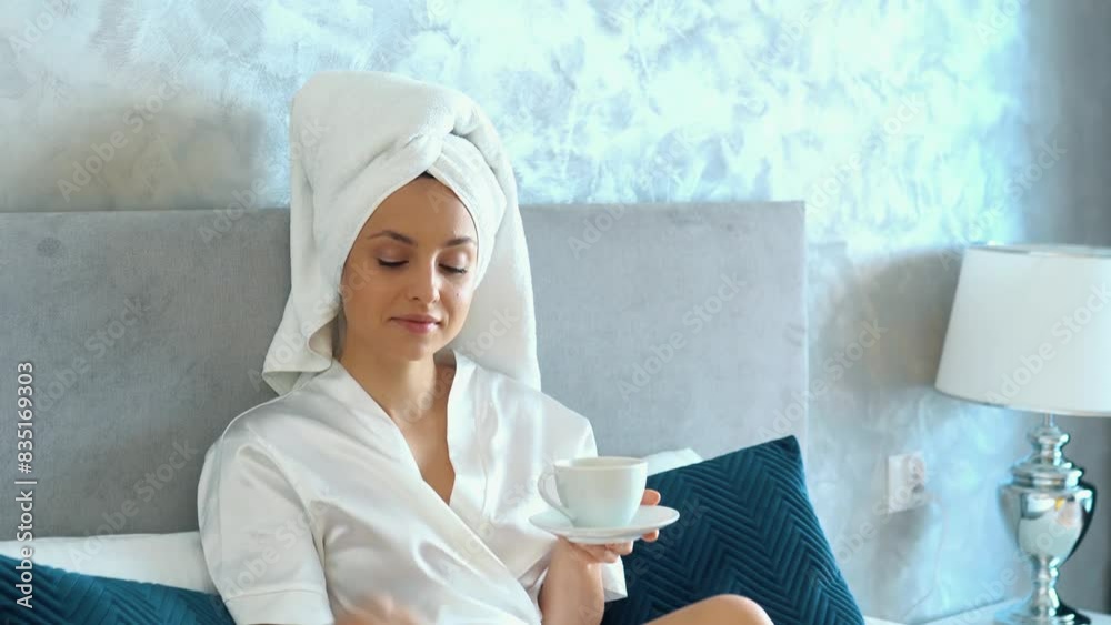 Young beautiful Caucasian woman is sitting in her bed at bedroom. Girl is wearing white bathrobe and towel on her head, she is drinking hot coffee from white cup, female is switching on tv. Young beautiful Caucasian woman is sitting in her bed at bedroom. Girl is wearing white bathrobe and towel on her head, she is drinking hot coffee from white cup, female is switching on tv.