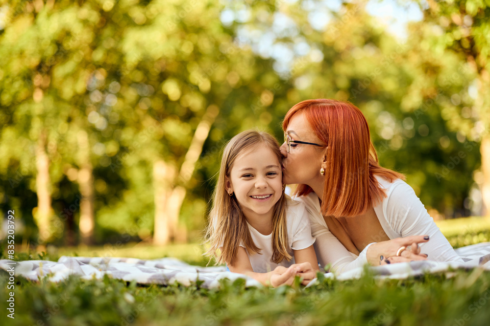 Lovely little girl smiling for the camera while her mother kisses her on the cheek, lying on a blanket at the park. Lovely little girl smiling for the camera while her mother kisses her on the cheek, lying on a blanket at the park.