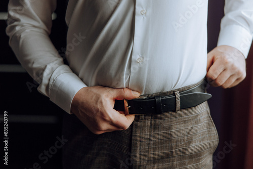 A man is adjusting his belt while wearing a white shirt. Concept of formality and attention to detail, as the man takes the time to ensure his belt is properly fastened