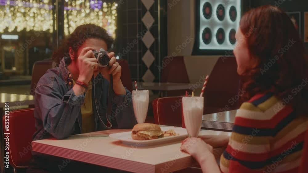 Back shot of happy Caucasian man taking photo for memory of his red haired sweetheart during date in neon lit retro styled cafe in evening
