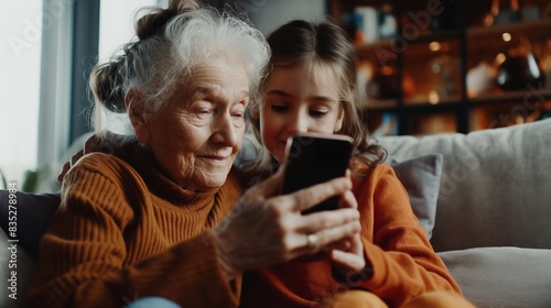 An elderly woman and a young girl are sitting on a sofa, closely looking at a smartphone together, sharing a bonding moment.