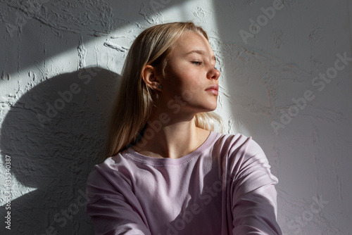 A pensive teenage girl in a pink jacket sits against a wall in the sunlight and looks to the side.