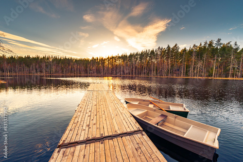 Small forest lake. Sunset behind the forest. Wooden bridge with boats. Small waves. View from the bridge. Summer day. View from the shore. Beautiful nature. Russia, Europe.