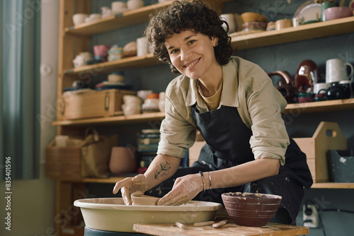 Portrait of cheerful female artisan wearing apron smiling at camera while shaping bowl on potters wheel