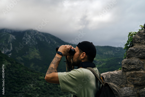 Travel and adventure concept. A young man with a retro-style green backpack stays in old town Bar in Montenegro country. Guy Looking at nature and green forest in mountains using binoculars.