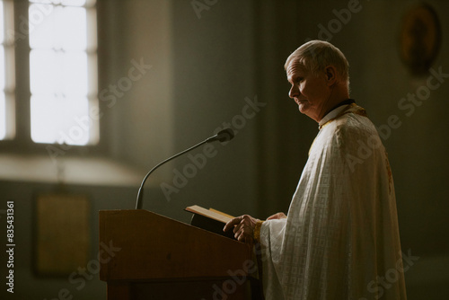 Senior Caucasian Catholic priest standing at lectern with mic opening Bible book, copy space