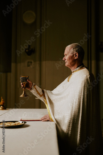Vertical side view shot of senior Caucasian Catholic priest holding cup wine conducting Holy Communion rite in church, copy space