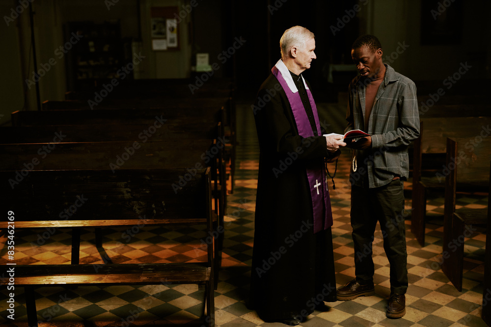 Obraz premium Long shot of ethnically diverse senior Catholic priest and young Black man standing indoors discussing religious text, copy space