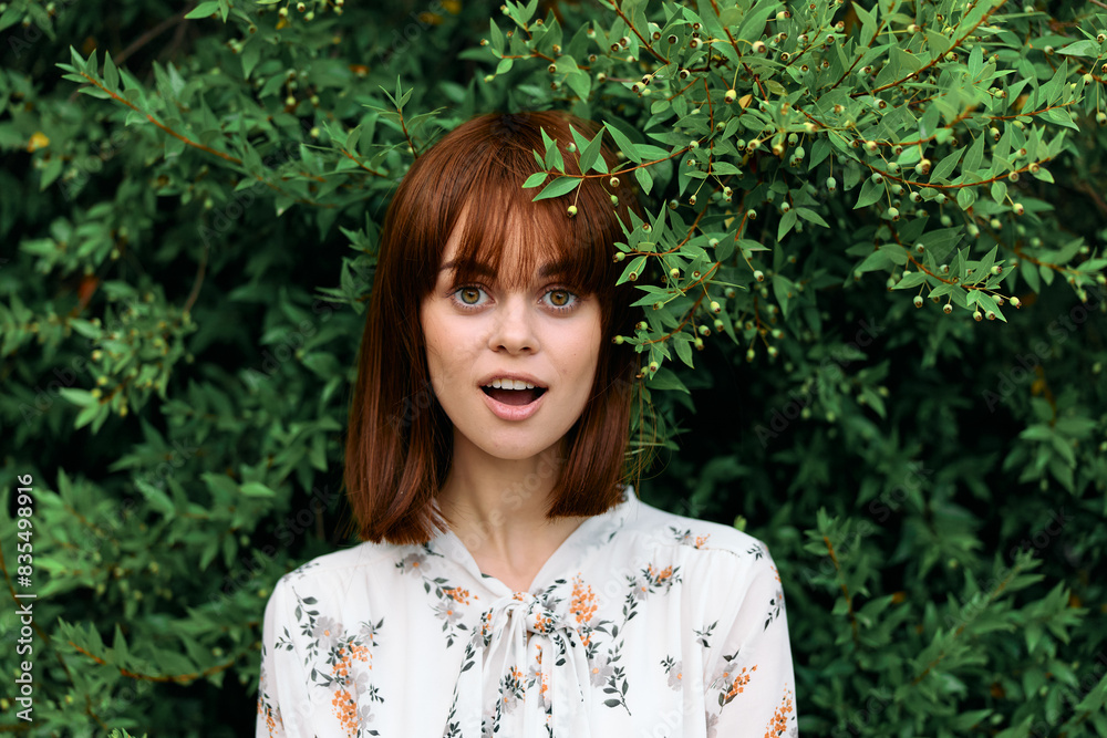 Vibrant redhaired woman posing gracefully in front of lush green leafy bush Vibrant redhaired woman posing gracefully in front of lush green leafy bush