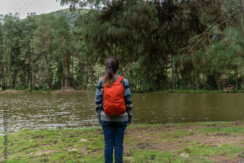 A young traveler standing in front of a pond in a pine forest.