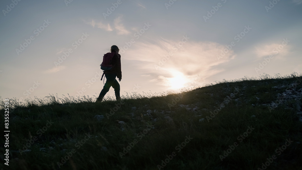 Female hiker silhouette with a backpack walking and following the trail uphill, enjoying the sky at sunset. Inspiration and healthy lifestyles concepts.