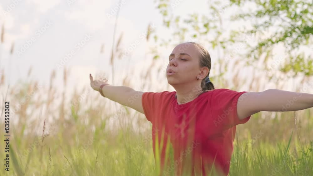 A woman wearing a red shirt practices yoga in a field of green grass. She is moving her arms in a fluid and graceful manner. The sun is shining and the weather is beautiful. A woman wearing a red shirt practices yoga in a field of green grass. She is moving her arms in a fluid and graceful manner. The sun is shining and the weather is beautiful.