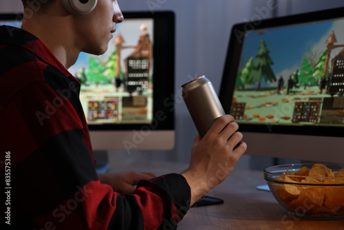Εκτύπωση καμβά Young man with energy drink and chips playing video game at wooden desk indoors,