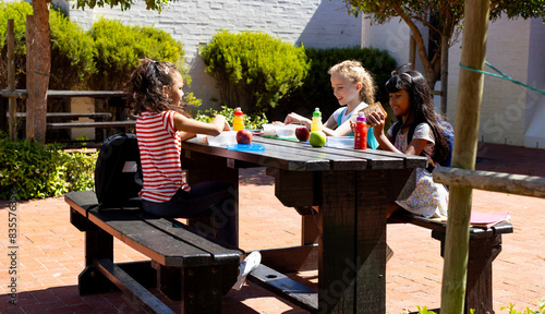 Three children enjoy a sunny outdoor lunch at a wooden table in school