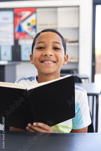 Biracial boy reading a book in a classroom at school, with copy space