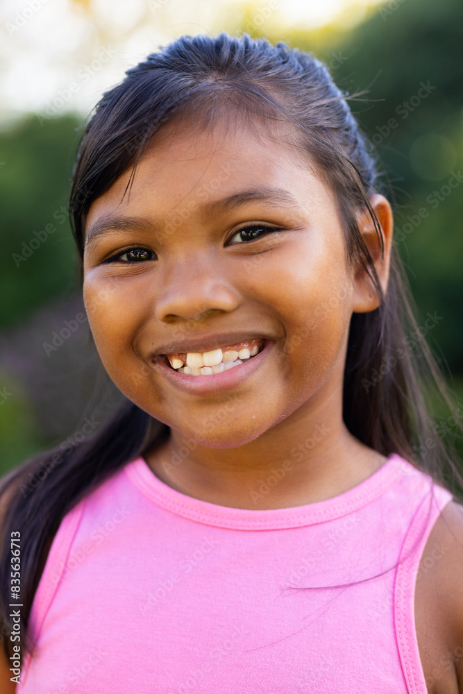 Outdoors, young biracial girl smiling brightly, wearing a pink top with greenery Outdoors, young biracial girl smiling brightly, wearing a pink top with greenery