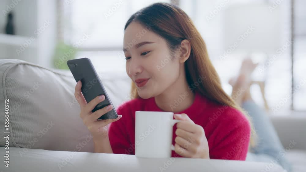 Attractive smiling young Asian woman using mobile phone while sitting on the sofa at home Attractive smiling young Asian woman using mobile phone while sitting on the sofa at home