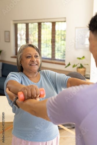 Biracial male physiotherapist helps senior female patient exercise with light weights at home photo