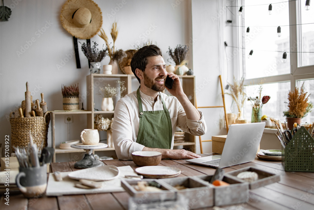 Successful businessman owner of shop made of clay talking to customer. Caucasian male potter entrepreneur talking to buyer of handmade bowl sitting at table and taking order using laptop. Successful businessman owner of shop made of clay talking to customer. Caucasian male potter entrepreneur talking to buyer of handmade bowl sitting at table and taking order using laptop.