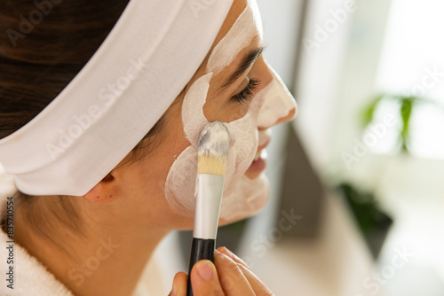 At home, young Caucasian woman applying facial mask with brush in modern home spa photo