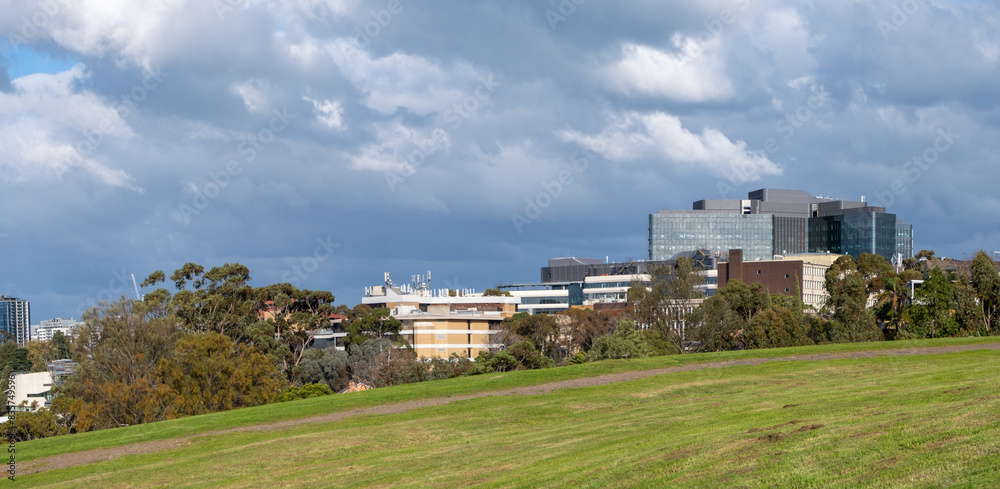 Panoramic background texture of a grass lawn on a hill with an urban modern buildings skyline and cloudy sky in the distance.Panorama of a green hillside outdoor ground near city of Maribyrnong Panoramic background texture of a grass lawn on a hill with an urban modern buildings skyline and cloudy sky in the distance.Panorama of a green hillside outdoor ground near city of Maribyrnong