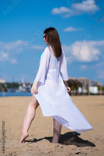 Beautiful, attractive, happy girl posing on a sandy beach on a sunny day in a blue swimsuit, sunglasses and a white beach tunic. Beachwear