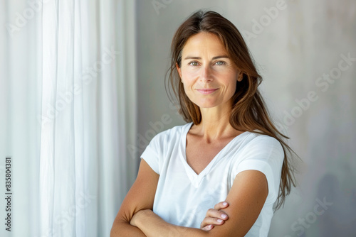 Portrait of a confident woman smiling, standing by a window with natural light. Free Download Portrait of a confident woman smiling, standing by a window with natural light. - GFXLoad