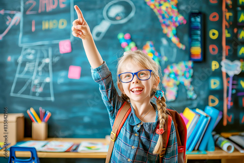 Enthusiastic young boy with glasses pointing up in a colorful, creative classroom with chalk drawings on the blackboard.