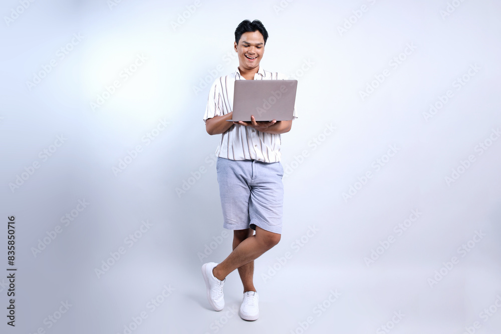 Full Length Of Happy Young Asian Man Standing Crossed Legs Wear Casual Style Using Laptop Isolated On White Background