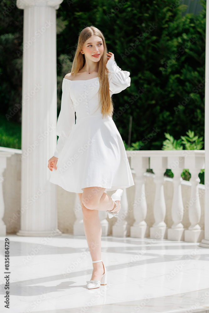 A woman in a white dress is standing in front of a white pillar. She is wearing white shoes and has long hair