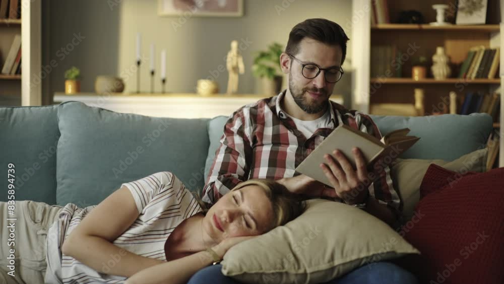 Slow-motion shot of a young caring husband reading story from book to her tired wife who is lying on her husband's lap on the sofa Slow-motion shot of a young caring husband reading story from book to her tired wife who is lying on her husband's lap on the sofa