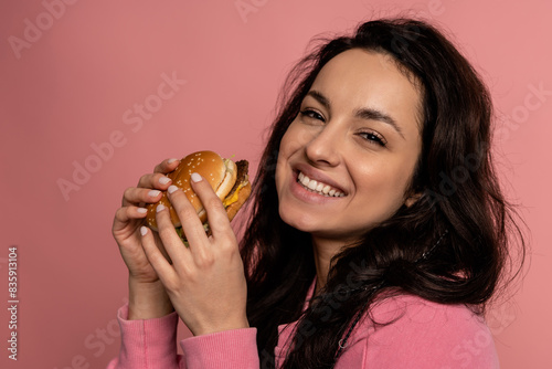 Pleased brunette with a delicious cheeseburger in her hands smiling during the studio photo shoot. Appetite and favorite fast food concept