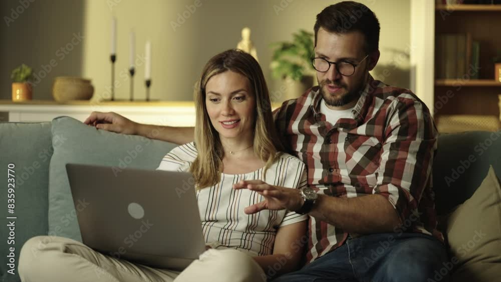Young business couple looking at their laptop screen and discussing business while sitting on couch in living room Young business couple looking at their laptop screen and discussing business while sitting on couch in living room