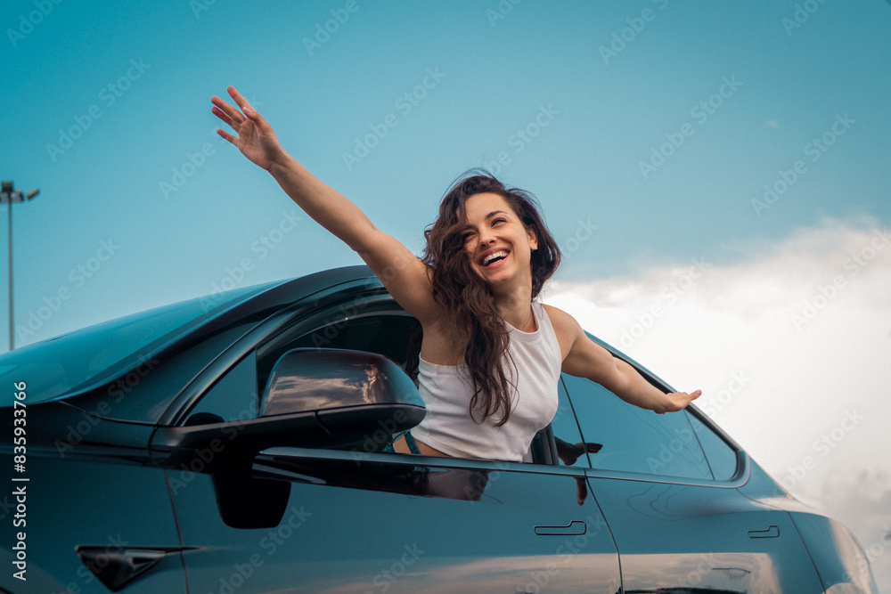 © Nataliya - Positive brunette lady enjoying travel while sticking out the side view window and holding hands to the side. Travel adventure drive, happy summer vacation concept
