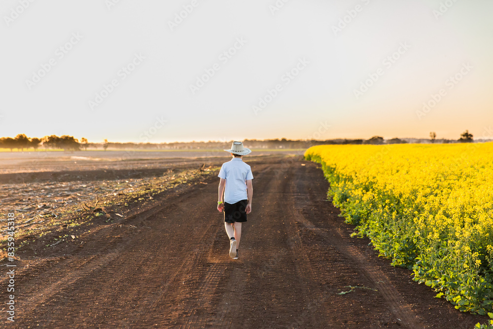 Boy wearing hat walking on dirt road next to canola paddock on farm Boy wearing hat walking on dirt road next to canola paddock on farm
