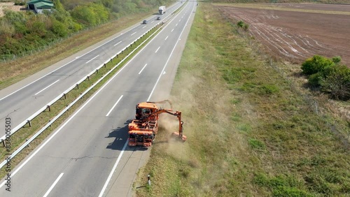 Top aerial view of a suburban highway with wheeled tractor lawnmover mowing the green grass on roadside at summer day