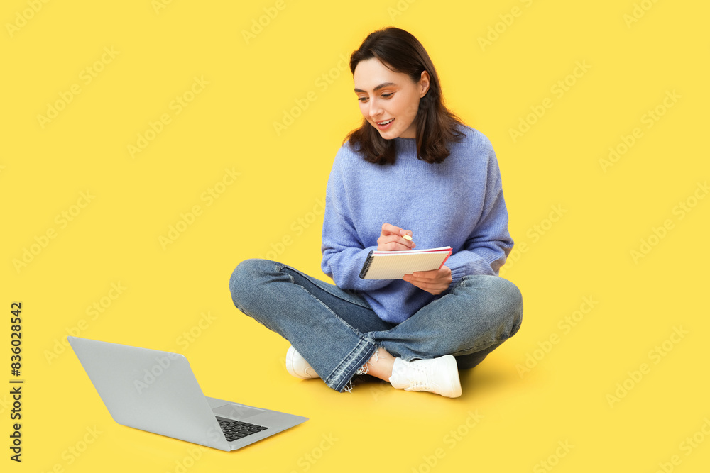 Pretty young woman working with laptop and notebook while sitting on yellow background