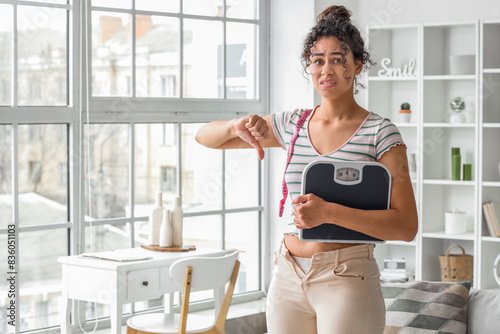 Upset African-American woman in tight pants with tape measure and weight scales showing thumb-down at home. Diet concept
