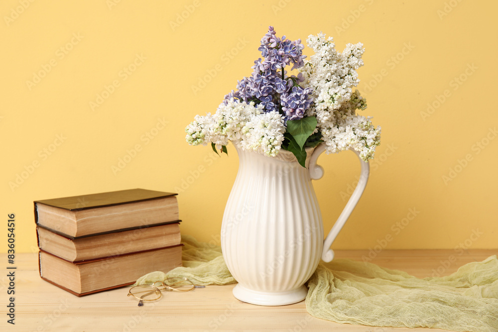 Jug with beautiful blossoming lilac and books on wooden table against yellow background Jug with beautiful blossoming lilac and books on wooden table against yellow background