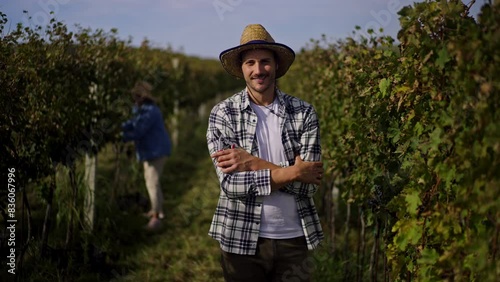 Young handsome farmer in hat standing in the vineyard, looking at camera and smiling. Two female winemakers ar standing in the background