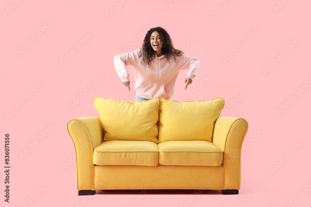 Young African-American woman pointing at yellow sofa on pink background Young African-American woman pointing at yellow sofa on pink background