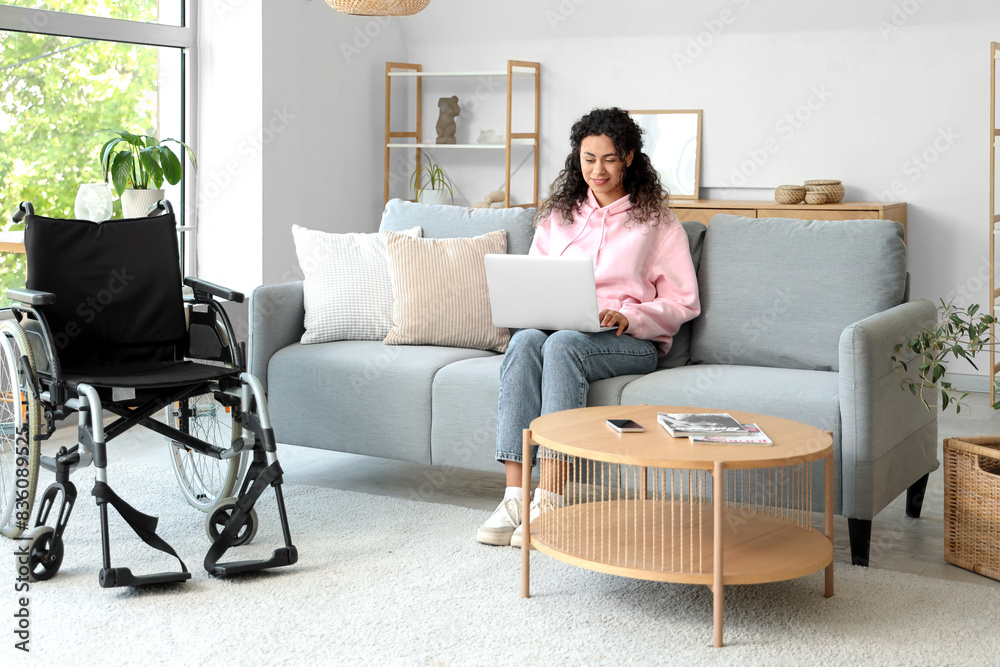 Young African-American woman with laptop and wheelchair sitting on sofa at home