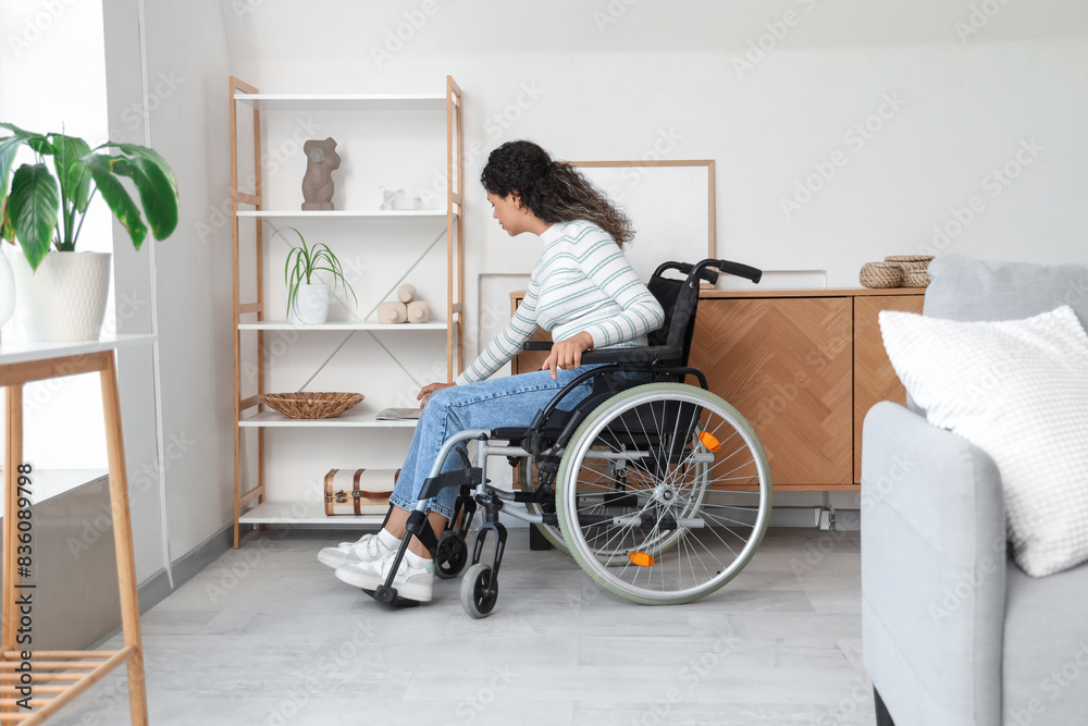 Young African-American woman in wheelchair taking magazine from shelf at home Young African-American woman in wheelchair taking magazine from shelf at home