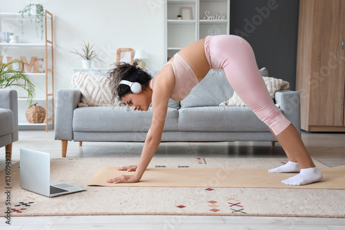 Sporty African-American woman with laptop and headphones exercising on yoga mat at home