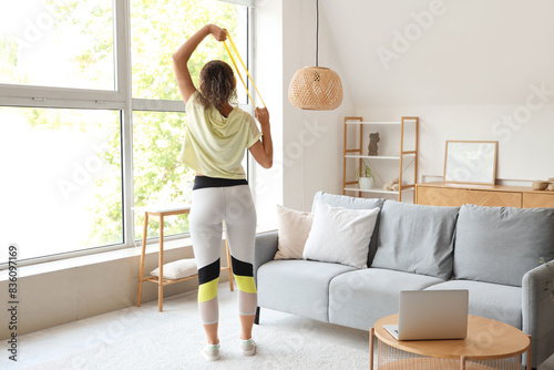 Sporty African-American woman with laptop and resistance band exercising at home photo