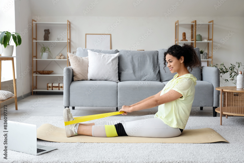 Sporty African-American woman with laptop and resistance band exercising on yoga mat at home Sporty African-American woman with laptop and resistance band exercising on yoga mat at home