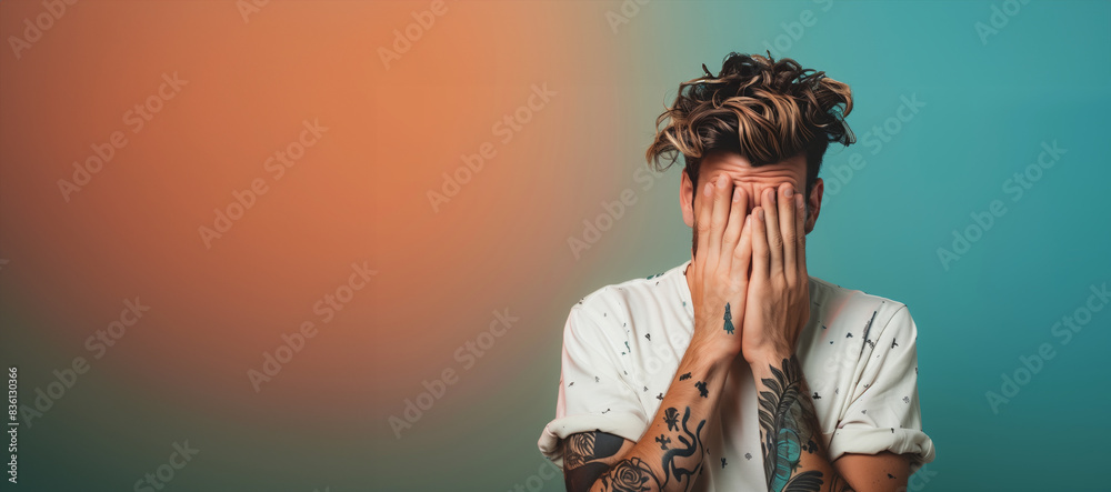 A man with tattoos on his arm. The background is a mix of orange and blue colors. Man's facial expression. a handsome italian man feeling disappointed and covers his face with both his hands A man with tattoos on his arm. The background is a mix of orange and blue colors. Man's facial expression. a handsome italian man feeling disappointed and covers his face with both his hands
