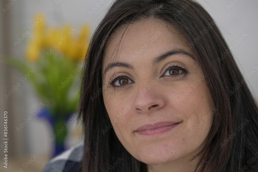 Obraz premium Woman with a thoughtful expression, close-up of face, subtle smile, dark hair, natural beauty, wearing a checkered shirt, indoor setting, blurred yellow flowers in background