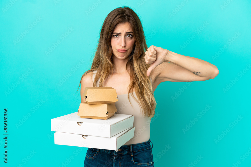 Caucasian woman holding pizzas and burger isolated on blue background showing thumb down with negative expression Caucasian woman holding pizzas and burger isolated on blue background showing thumb down with negative expression